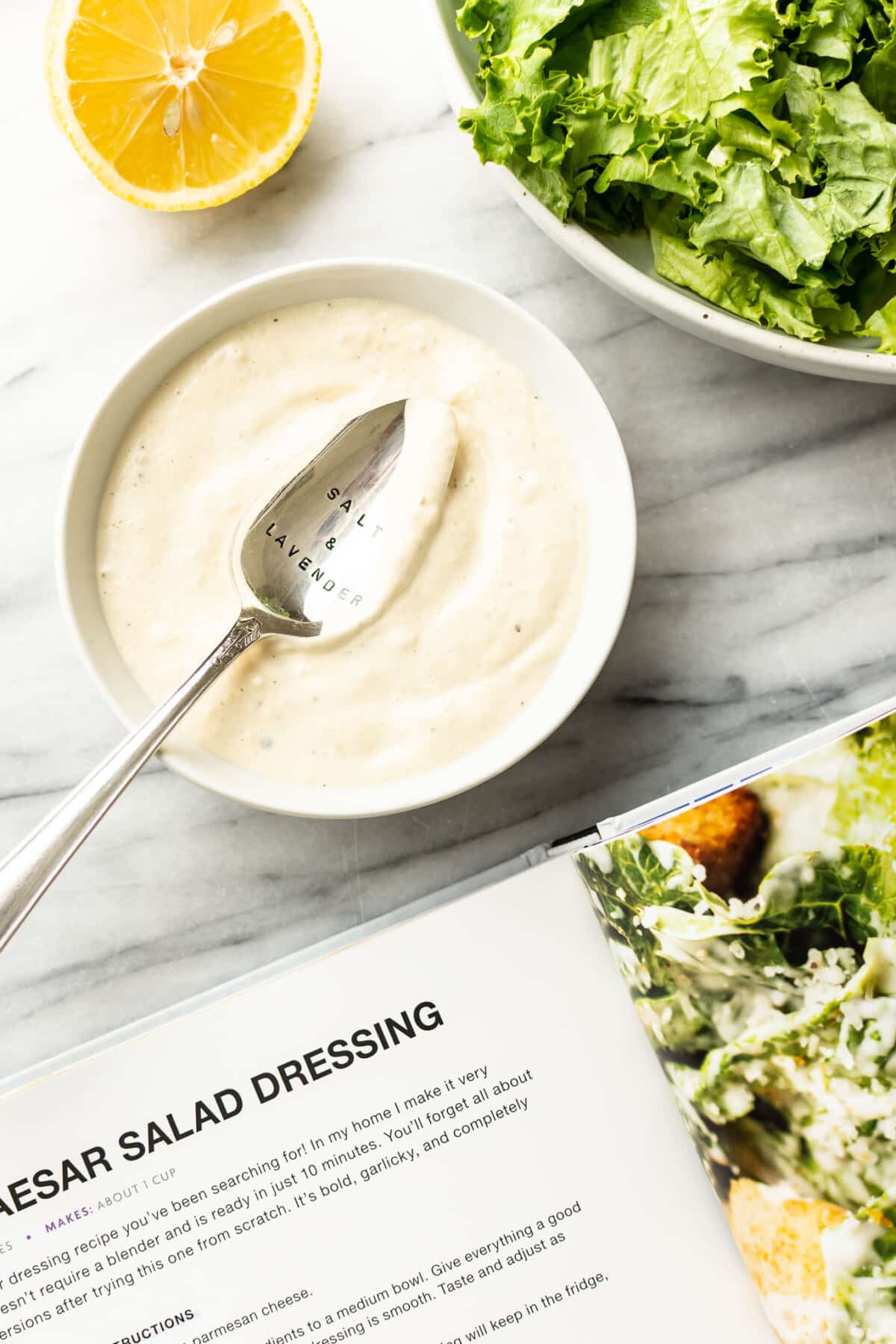 a bowl of homemade caesar salad dressing next to lettuce and a salt and lavender cookbook