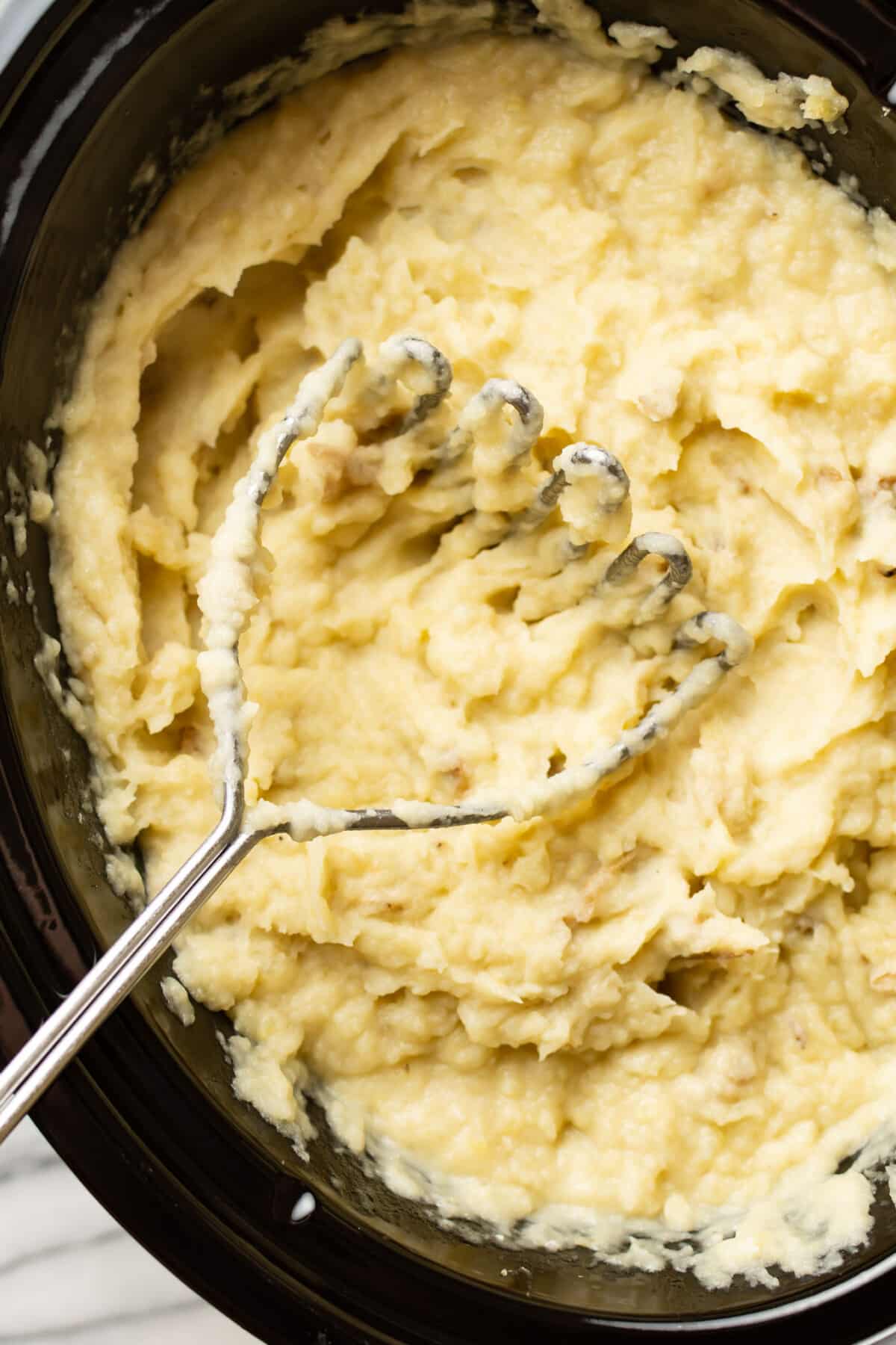 a slow cooker with mashed potatoes and a handheld masher