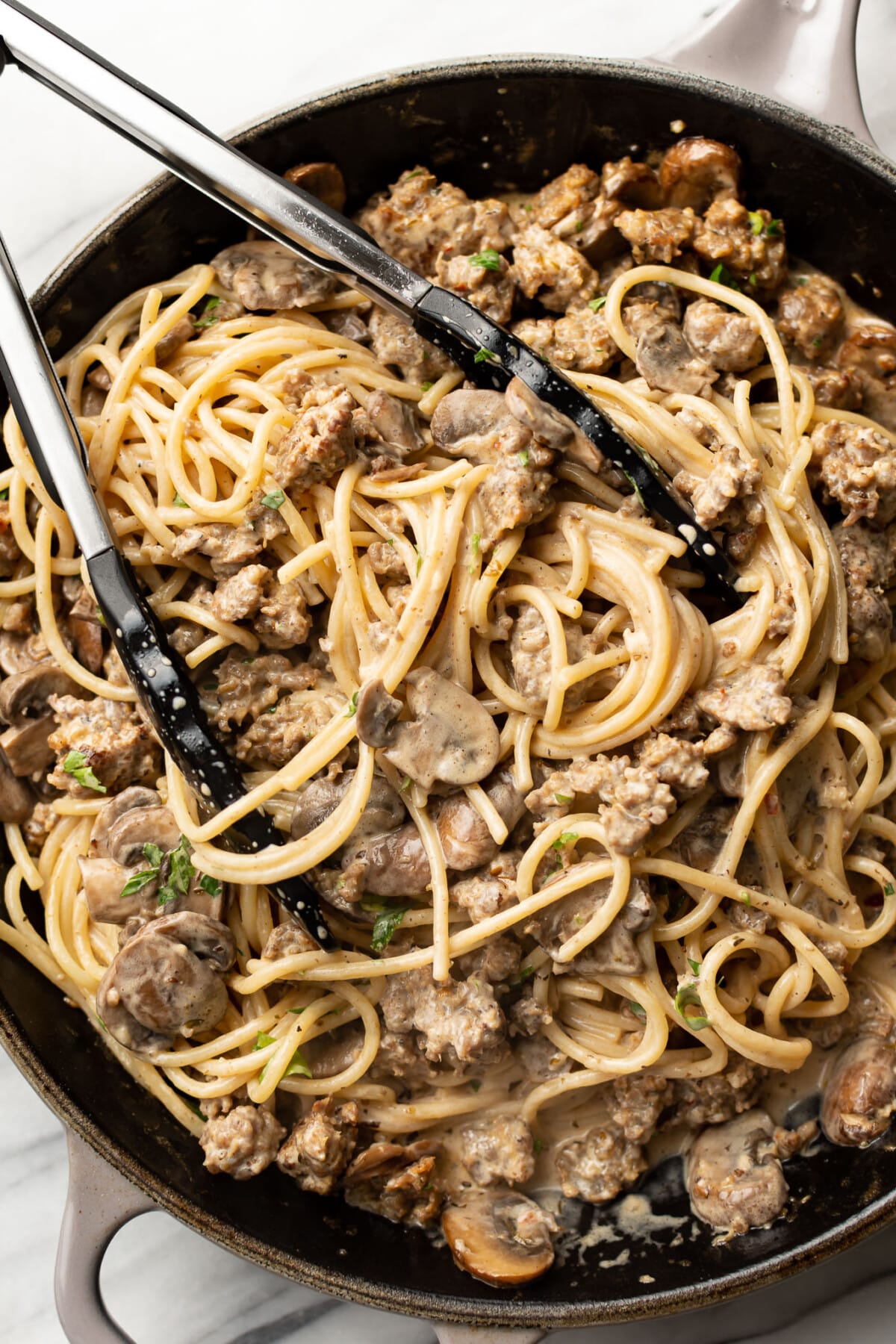 a skillet with sausage and mushroom pasta and kitchen tongs
