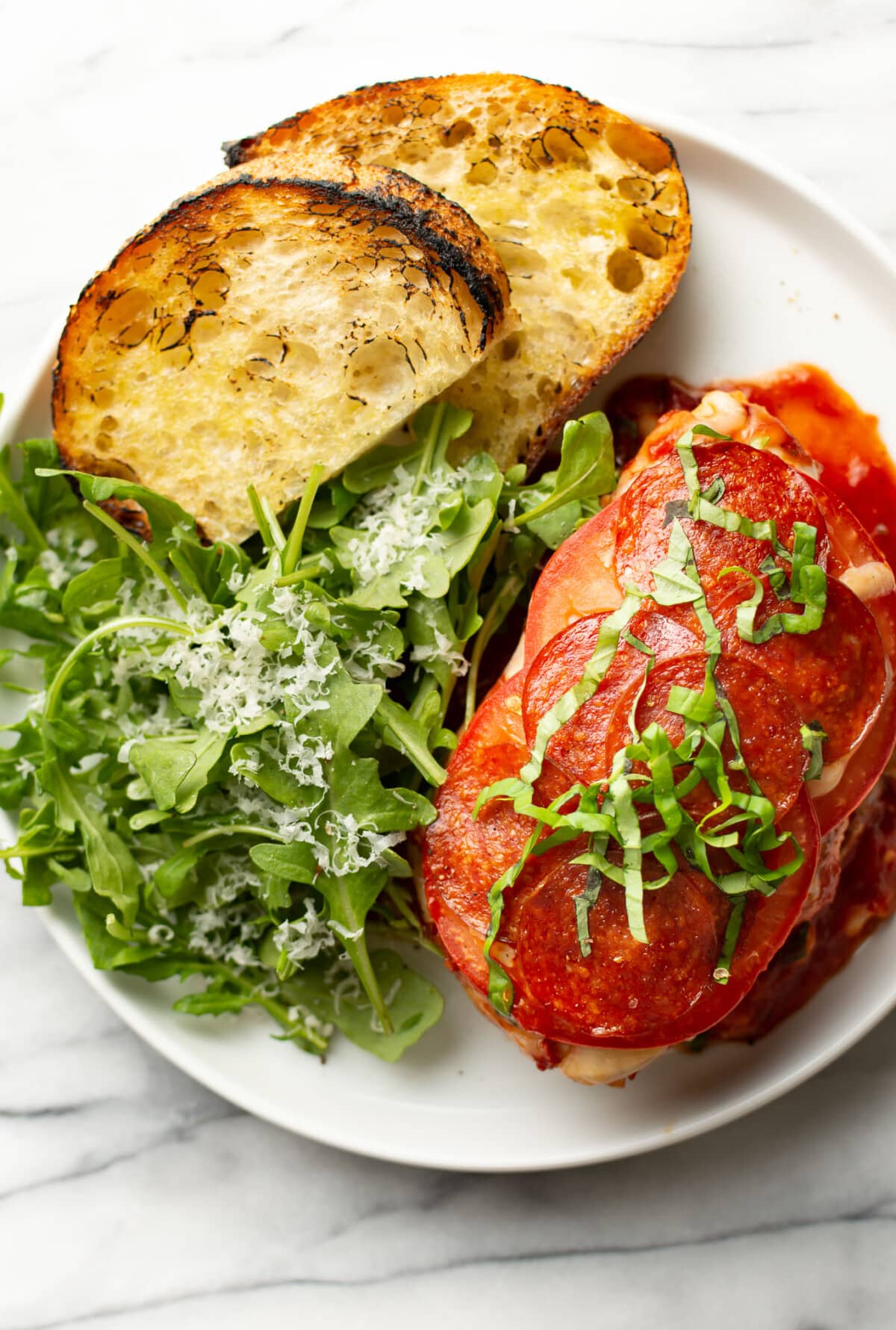 a plate with pizza chicken, toasted bread, and arugula salad