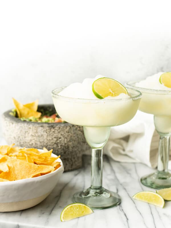 two frozen margaritas next to a bowl of guacamole and chips